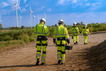 A team of engineers in high visibility safety gear stand together at a wind farm, conducting...