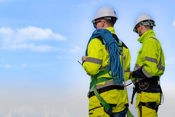 Two engineers in high visibility jackets and helmets are inspecting a wind farm. One is holding a mobile device, while the other is equipped with ropes for climbing, with turbines.