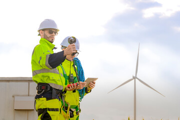 Two engineers wearing high visibility safety jackets and helmets measure wind speed with an...
