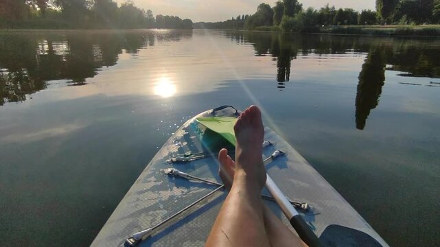 Bare legs of woman on SUP board on surface of river or lake with sun reflecting and with trees on banks on warm summer evening. Person swimming on SUP board on pond on calm day. Tourism rest sport