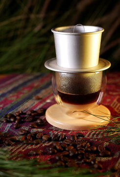 Vietnamese drip coffee in a glass with a metal filter on a wooden coaster, surrounded by coffee beans. The background features bokeh lights and green leaves, providing a calm and natural ambiance.