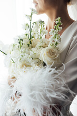 The morning of the bride, a woman wearing a white silk robe holds a wedding bouquet. Close-up photo.