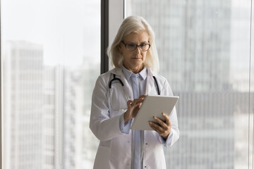 Serious older female doctor cardiologist look on tablet screen read virtual history case of...