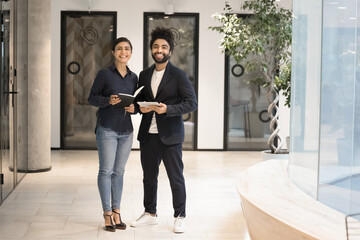 Two multiethnic happy successful business partners standing in office hall interior, smiling, looking at camera, holding notebook, tablet. Indian and Arab startup employees full length portrait