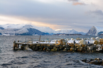 A wooden dock juts out into the water, surrounded by large rocks. The water is choppy and reflects the blue sky above.