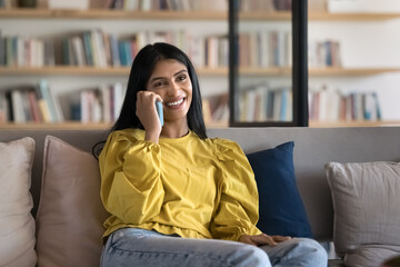 Obraz premium Cheerful attractive young Indian student woman speaking on cell in library, looking away, smiling, laughing, talking on mobile phone, enjoying conversation, wireless communication, telephone call
