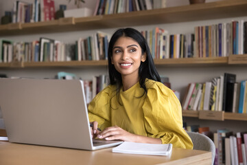 Positive successful Indian student girl woman working on research study in public library, typing on laptop, looking away, smiling, thinking on education, creative idea for essay, article