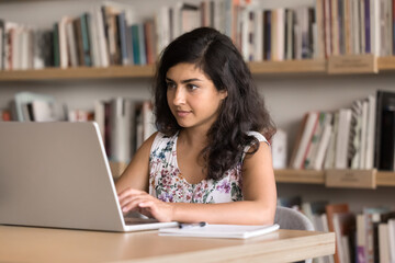 Focused young adult Indian student woman typing on laptop in public library, working on job project at table with bookshelves in background, reading professional information on screen