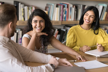 Happy young adult Indian student girls talking to teacher in library, laughing, sitting at meeting table with bookshelves in background. Cheerful business colleagues brainstorming, having fun