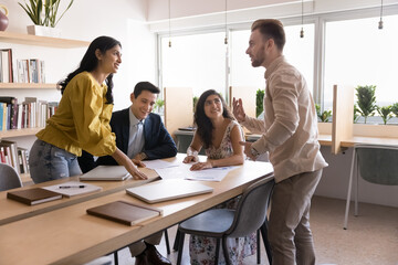 Positive multiethnic couple of young business colleagues discussing marketing reports, standing at large table, meeting with diverse team of colleagues, brainstorming, cooperating on project