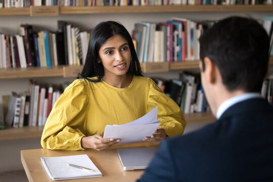 Young Indian recruit manager woman talking to job candidate at interview, holding paper resume. Diverse student and teacher meeting in library, discussing research study