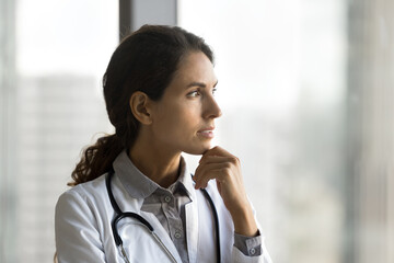 Fototapeta premium Headshot pensive millennial lady doctor general practitioner look at window ponder on treatment tactic in difficult clinical case. Thoughtful female family therapist reflect on medical ethics question