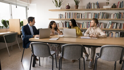 Diverse team of adult students talking at large table in library with bookshelves in background,...
