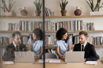Multiethnic couple of diverse business colleagues getting professional knowledge in library, using laptop together, discussing training course, online learning program. Candid shot with copy space