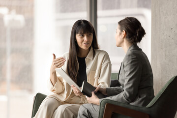 Two female office friends chatting in office lobby on work break, discussing online project,...