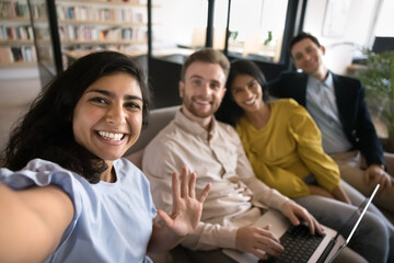 Happy young Indian manager girl in casual taking selfie with office friends, business colleagues sitting on couch on work break, having fun, laughing, waving hand hello at camera