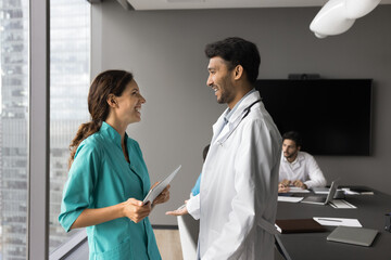 Friendly diverse young doctors general practitioners teammates stand at modern clinic office talk laugh. Multiethnic millennial medic workers physician and nurse chat discuss work case share good news