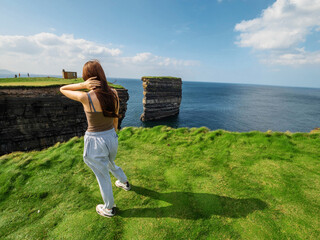 Teenager girl standing by edge of a cliff and looking at stunning Dun Briste sea stack and blue...