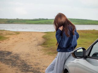 A woman is sitting on the side of the road next to a silver car. She is wearing a blue hoodie and gray sweatpants. The scene is quiet and peaceful. Model looking at calm nature scene. Travel to nature