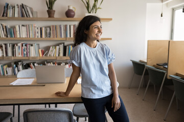 Cheerful young Indian entrepreneur woman standing at workplace table in co-working, looking away,...