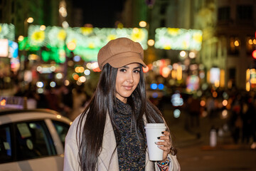 Obraz premium Beautiful young woman is holding a coffee cup on a blurred night city street with lights