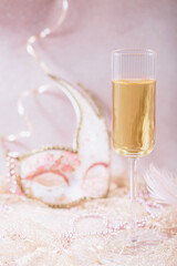 Masquerade ball holiday background. Glass of white champagne, Venetian carnival mask and feathers on light pink background