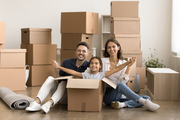 Starting new life. Happy homeowners posing for lifestyle family portrait sit on floor with belongings packages at moving day. Smiling mom dad hold hands of playful kid looking at camera from big box