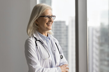 Candid smiling mature female doctor modern private medical center leader stand by window look at distance make ambitious plans visualize successful work feel happy for perfect care of patients health