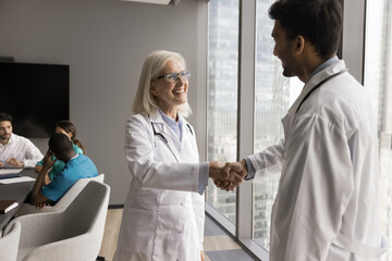 Friendly senior female chief physician shake hand to millennial male medic get acquainted with new teammate. Mature woman head doctor of clinic department meet young specialist glad to welcome in team