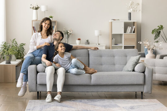 Happy homeowners. Friendly family married couple preteen age daughter spend time together cuddling on couch at cozy living room talk laugh enjoy being renters tenants of modern apartment. Copy space