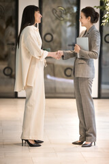 Happy friendly confident businesswomen meeting in office hallway, greeting with handshakes, talking, smiling, saying hello, goodbye. Female business leader congratulating colleague, shaking hand