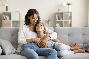 Tender moments. Smiling little girl clings closer to beloved single mom sitting on couch feeling...