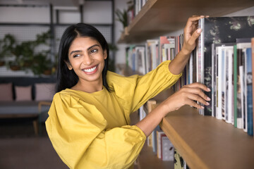 Cheerful young Indian student girl enjoying reading hobby, choosing book in library, bookstore, taking textbook from bookshelf, looking away, smiling, laughing, looking for information