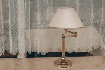 A silver lamp with a white shade sits on a table in front of a lace curtain
