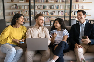 Happy diverse group of friends holding gadgets, talking and laughing on couch, resting on sofa in modern co-working office lobby, enjoying online and offline communication