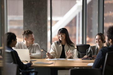 Cheerful Latin female team leader holding group meeting, negotiation, jog interview. Business mentor, teacher, coach talking to interns, laughing, joking at conference table
