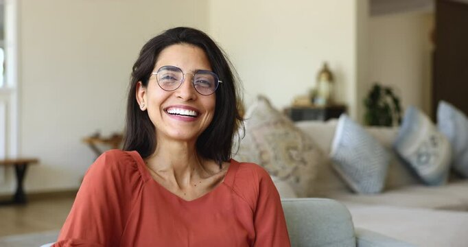 Close up front face view, head shot happy young 30s Brazilian woman wear eyeglasses having candid white-toothed smile look at camera seated on armchair in living room. Homeowner portrait, home pastime