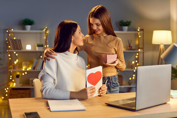 Smiling cute teen girl child congratulating her happy young mother on holiday. Daughter giving her mother sitting at the desk at home present gift box and greeting card. Mother's day concept.
