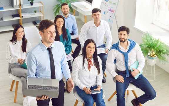 Businessman surrounded by colleagues engages in a group office meeting, participating by asking a question. This scene epitomizes teamwork and effective communication in a business environment.