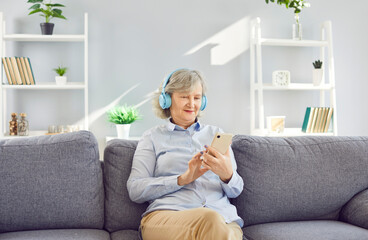 Senior smiling relaxed woman sitting on sofa at home and listening to music with headphones and...