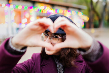 Young smiling lovely woman showing shape heart with hands and looking through it standing outside over blurred colored lights background on winter. Focus on face
