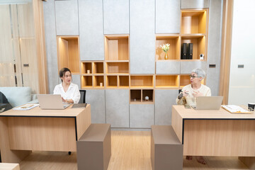 A woman wearing glasses is sitting at a desk with a laptop and a cell phone. She is looking at the...