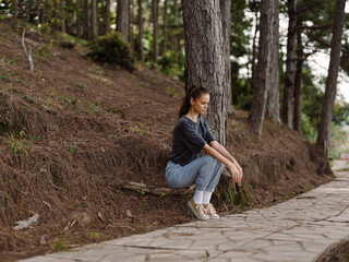 Thoughtful young woman sitting on the ground against a tree in a serene forest, reflecting in nature s calm surroundings Relaxation and mindfulness concept