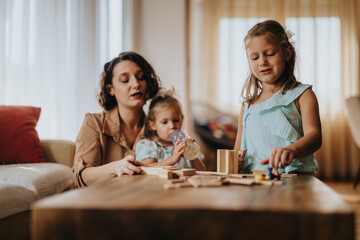 A mother spends quality time with her two daughters, playing and building creatively on a wooden table. The scene reflects bonding, learning, and family happiness in a cozy home environment.