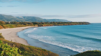 A serene beach view with gentle waves, lush greenery, and mountains in the background under a clear blue sky.