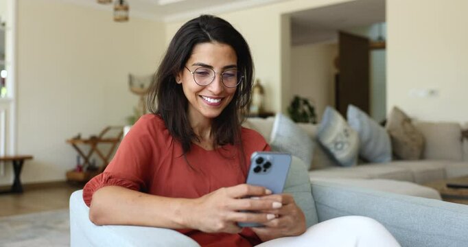 Brazilian woman relaxing seated on cozy armchair with smartphone, enjoy remote chat with friend, using new mobile e-dating application for singles, check messages, responding on sms on weekend at home