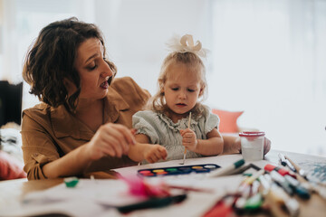 A mother and her young daughter enjoy a craft activity together at home. The scene depicts bonding through creative play and learning with paints and supplies on the table.