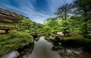 Obraz premium A serene Japanese garden with a pond, a wooden bridge, and lush greenery under a partly cloudy sky.