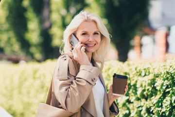 Portrait of charming lovely pensioner lady talk phone coffee spring good mood wear beige coat enjoy...
