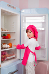 Little beautiful girl in a chef costume takes food from the refrigerator for cooking in the kitchen.	